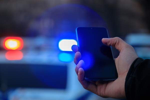 A hand holding a smartphone with blurred blue and red police lights in the background.