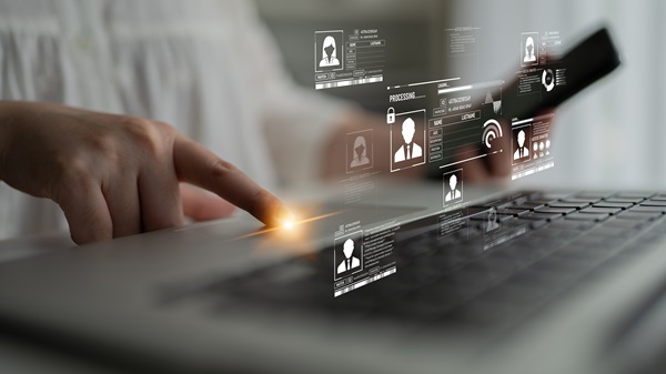 A person's hand typing on a laptop with digital profile icons and data fields floating above the keyboard, representing online identity and processing.