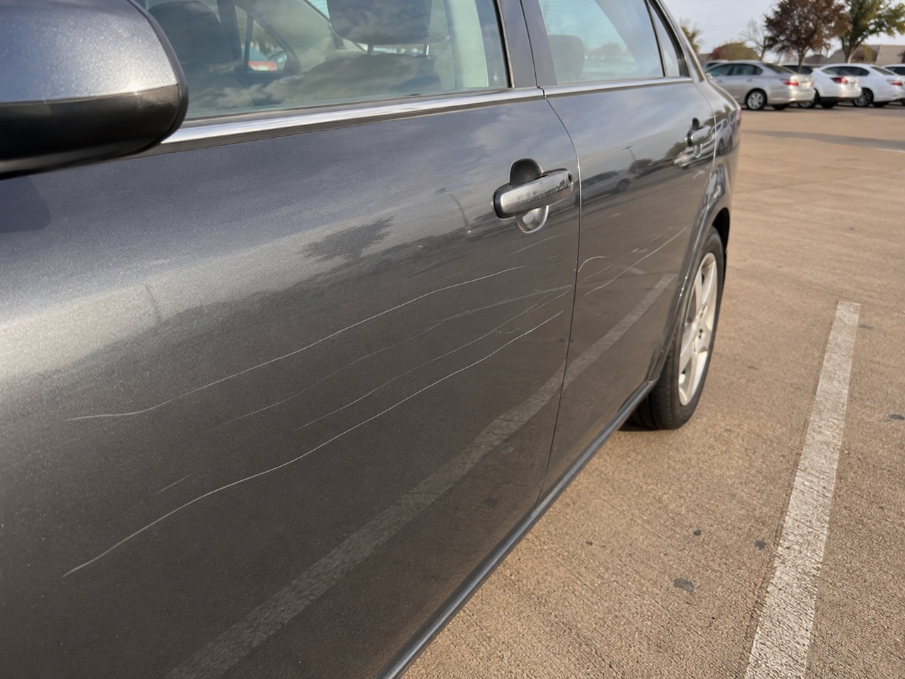 A close-up of a gray sedan door showing multiple deep scratches across the paint in a Texas parking lot, depicting intentional property damage that could result in criminal mischief charges and felony penalties under the Texas Penal Code.